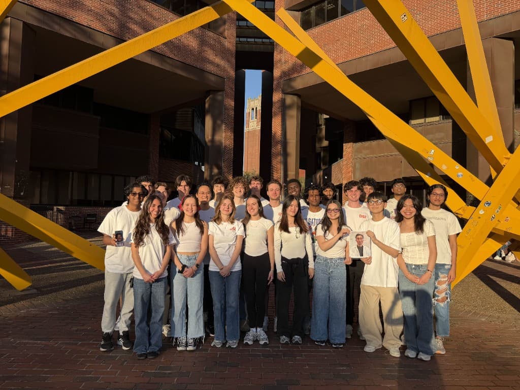About thirty DSI members in white shirts standing under the yellow steel sculpture outside Marston Science Library, Century Tower behind them